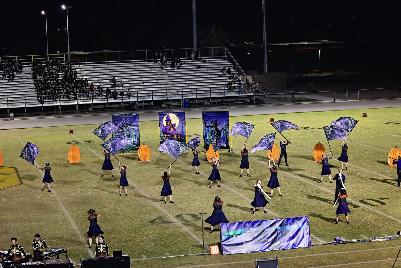 Color guard performing with spiderweb flags in front of a haunted house backdrop, with lit pumpkins across the field and the drumline in skeleton uniforms